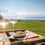 Auf einer Obstkiste stehen zwei Gläser Wein und ein belegtes Brot, im Hintergrund eine Wiese und der Bodensee